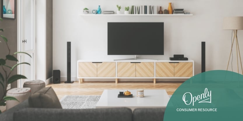 A tv is atop a tv stand behind a coffee table and underneath a bookshelf. 