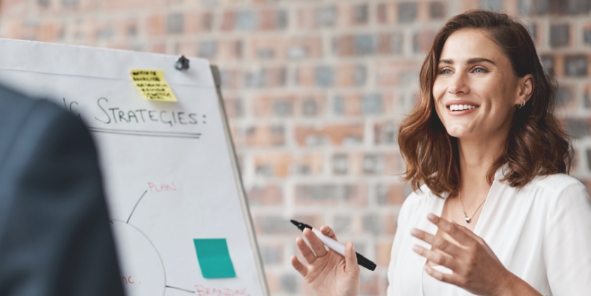 A woman smiles while holding a pen in front of a board outlining strategies in an office. 