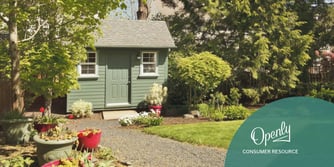 A small green building with two windows on either side of the door sits at the end of a path in a green yard. 