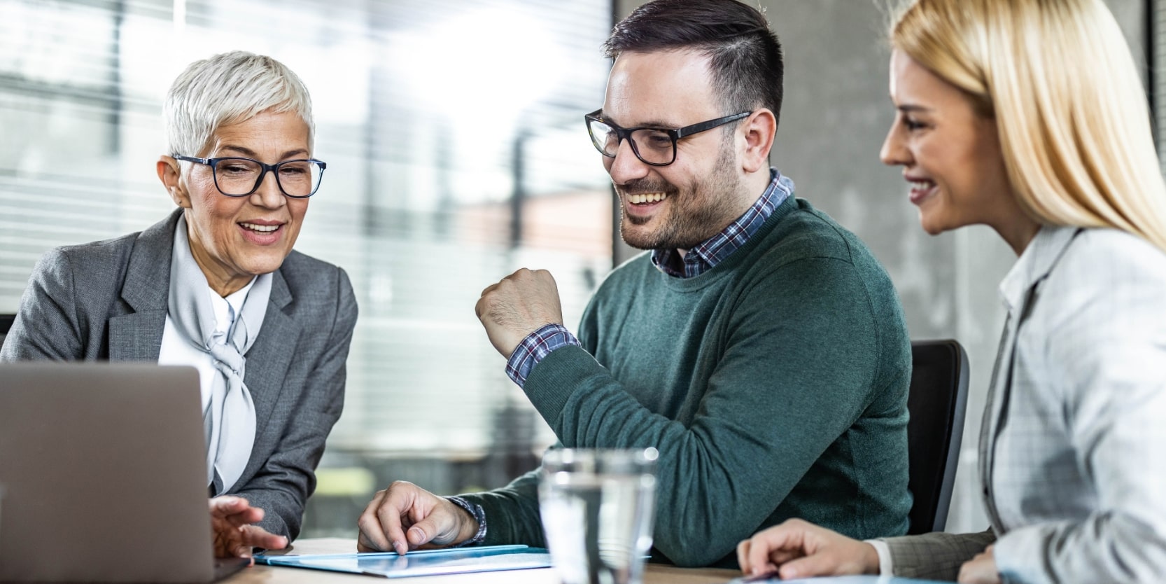A smiling businesswoman shows two smiling clients a laptop. 