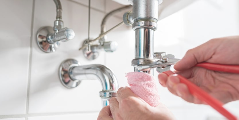 A person works on under-sink pipes 