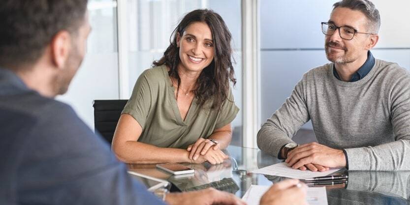 A man and woman sit at an office desk smiling at another man.