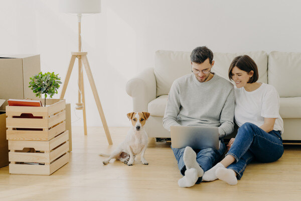 A couple sits on the floor with a dog, using a laptop, creating a relaxed atmosphere in their home