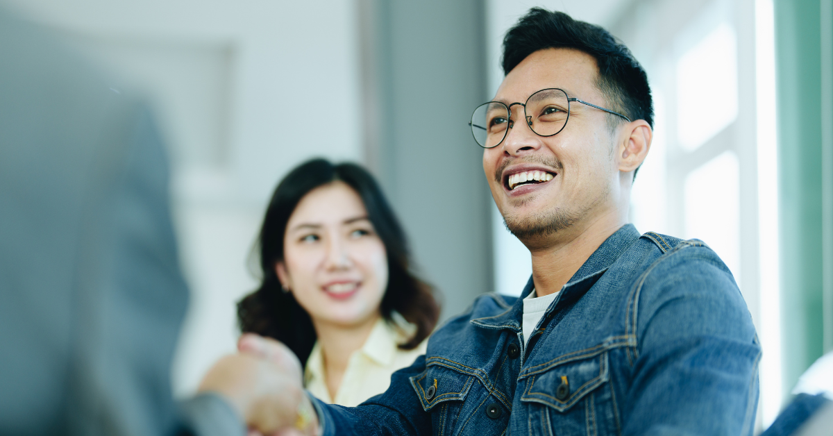 A smiling man shakes a business person's hand while a woman looks on. 