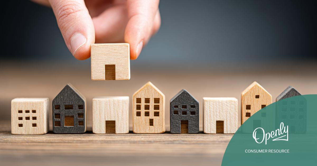 A series of wooden toy houses are lined up in a row on a table and a person holds an identical house above one of them.