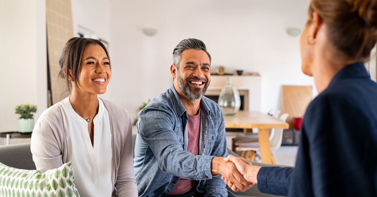 A smiling man shakes a woman's hand while a smiling woman sits on a couch next to him. 